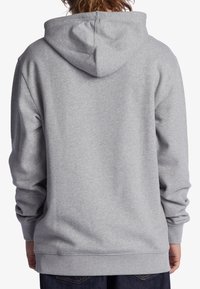 Persoon gekleed in een effen lichtgrijze capuchonsweater gezien van achteren, gecombineerd met donkerblauwe jeans.