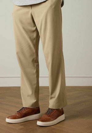 Beige trousers made of smooth fabric, tapered fit, paired with brown leather sneakers featuring white soles and dark laces on a wooden floor.