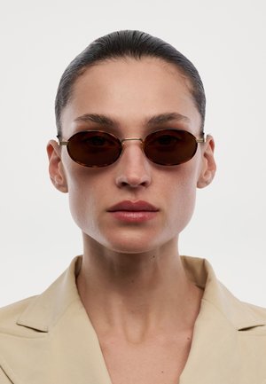 Person med bagstrøget hår iført ovale solbriller med skildpaddemønster og en beige jakke med krave, der vender fremad mod en hvid baggrund.