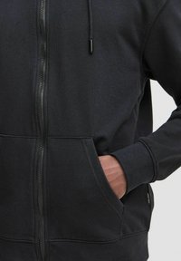 Black zip-up hoodie made of cotton blend fabric. Features kangaroo pocket, drawstring hood, and ribbed cuffs. Smooth texture with clean seams.