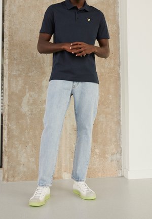 Marineblaues Poloshirt mit gelbem Logo, kombiniert mit hellblauen Jeans und weißen Turnschuhen mit grüner Sohle. Glatter Stoff und schlanke Passform.