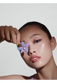 Gros plan d'un visage à la peau claire, lèvres brillantes et maquillage des yeux doux. Une délicate fleur violette est tenue près d'un œil, sur un fond neutre.