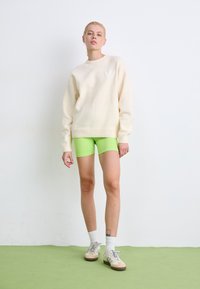 Crème oversized sweatshirt met een textuurafwerking, gecombineerd met aansluitende neon groene shorts en sneakers in neutrale tonen. Eenvoudige witte achtergrond.
