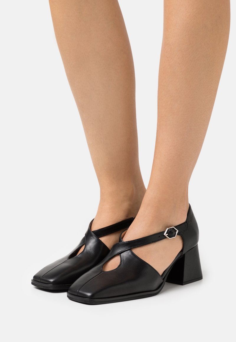 Chaussures en cuir noir avec un bout carré, des découpes et un talon épais. Dotées d'une bride de cheville réglable avec une boucle hexagonale en accent.