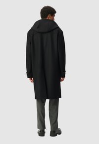 Cappotto lungo nero con cappuccio, caratterizzato da una linea dritta e un tessuto testurizzato. Indossato sopra pantaloni grigi a righe sottili e scarpe nere.