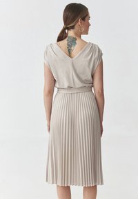 Abito plissettato beige con un profondo scollo a V drappeggiato nella parte posteriore e maniche a cappuccio. Presenta una cintura in vita e un tessuto liscio e strutturato.