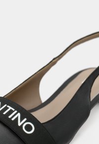 Zapato slingback negro hecho de cuero elegante, con una amplia banda elástica con letras blancas. Diseño suave y minimalista con punta puntiaguda.