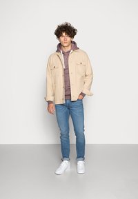 Giacca beige con due tasche frontali, indossata sopra un hoodie bordeaux con una cerniera bianca, abbinata a jeans blu e sneakers bianche.