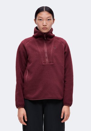 Femme portant un pull polaire bordeaux avec un col montant et une fermeture éclair trois quarts, debout devant un fond clair uni.