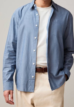Homme portant une chemise bleu à fines rayures déboutonnée sur un t-shirt blanc, pantalon beige et ceinture en cuir marron, avec une main dans la poche.