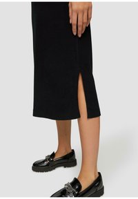 Robe en tricot noire avec une fente sur le côté et un ourlet côtelé, associée à des mocassins noirs brillants avec des détails en métal décoratifs.