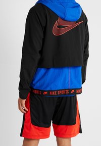 Person som bär en svart och blå Nike-jacka med en stor röd logotyp på ryggen samt svarta och röda träningsshorts, vänd bort.