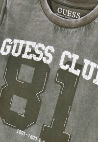 Pilkos organinės medvilnės marškinėliai, su įtrūkusiomis baltomis raidėmis "GUESS CLUB" ir skaičiumi "81", su baltais siuvinėjimo akcentais.