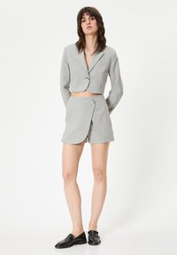 Blazer grigio corto con chiusura a un bottone e pantaloni corti avvolgenti grigi coordinati. Tessuto liscio con minima texture, abbinato a scarpe eleganti nere.