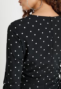 Černé šaty s bílými puntíky, s kulatým výstřihem a viditelným zipem na zádech. Materiál je hladký s mírným leskem.