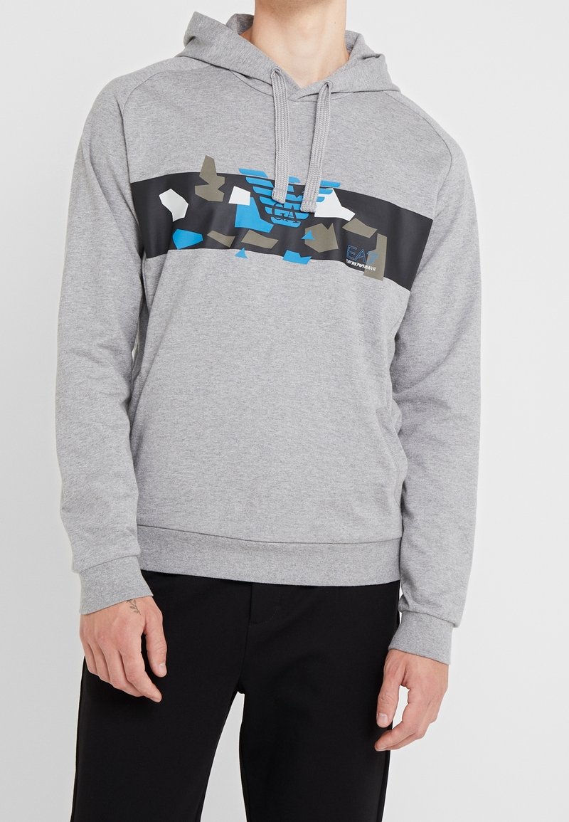 Personne portant un sweat à capuche gris avec une bande de logo abstrait bleu et noir sur la poitrine, assorti à un pantalon noir, debout sur un fond blanc.