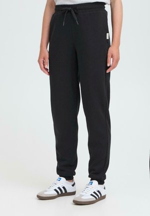 Person som bär svarta joggers med dragsko och vita Adidas-sneakers med svarta ränder, som står mot en enkel ljus bakgrund.