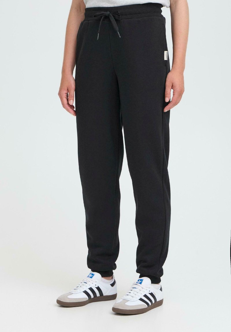 Person som har på seg svarte joggebukser med snøring og hvite Adidas joggesko med svarte striper, står foran en ensfarget lys bakgrunn.