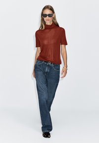 Rotes kurzärmeliges Oberteil mit Stehkragen, kombiniert mit lockeren dunkelblauen Jeans und schwarzen Sandalen mit offenen Zehen, Modell läuft geradeaus.