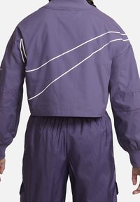 Purple cropped jacket with white diagonal stripes on the back, featuring a ribbed collar and oversized sleeves, paired with textured pants.