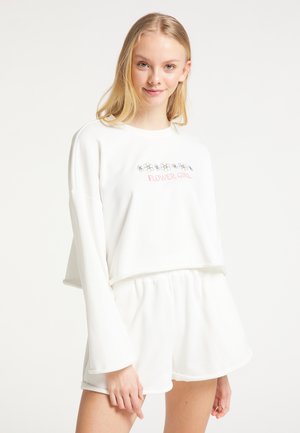 Junge Frau trägt ein weißes Langarmshirt mit dem Schriftzug "FLOWER GIRL" und Blumenmotiven, kombiniert mit passenden weißen Shorts.