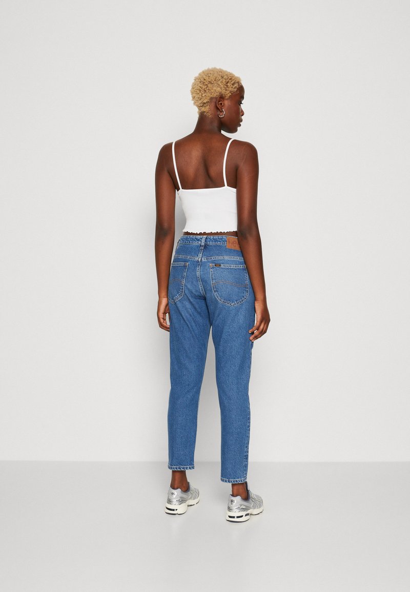 Weißes, kurzes Tanktop und blaue, hoch taillierte Jeans mit lockerer Passform, fransigem Saum und heller Waschung. Silberne Sneakers runden den Look ab.