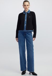 Zwarte geribbelde cardigan met een denim kraag en voorkant, voorzien van een logo patch en zilveren knopen. Gecombineerd met wijde blauwe jeans.