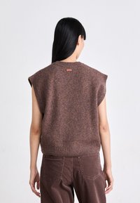 Ärmelloses Strickoberteil in einer Mischung aus Braun und Lila, mit einem gerippten Saum und einem kleinen Label auf der Rückseite. Getragen mit dunkelbraunen Hosen.