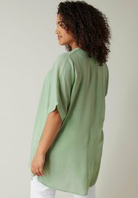 Ljusgrön, löst sittande blus med en slät, silkeslen textur, korta ärmar och en rundad nederkant. Har en subtil söm i ryggen.