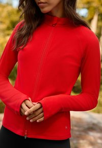Red zip-up jacket made of lightweight fabric, featuring long sleeves with thumbholes and a high collar. Smooth, stretchy texture.