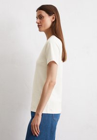 Helles Creme-farbenes T-Shirt mit kurzen Ärmeln und rundem Ausschnitt, das eine taillierte Form und eine glatte Baumwolltextur aufweist, kombiniert mit blauen Jeans.