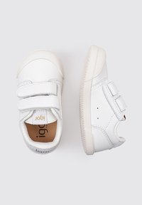 Zapatillas blancas para niños con dos correas de velcro, punta redonda, suela de goma texturizada y forro suave. Logo visible en la lengüeta.