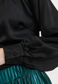 Black satin blouse with puffed sleeves and buttoned cuff, paired with a shiny green pleated skirt. Smooth texture and elegant design details.