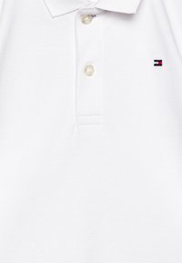 Polo shirt blanc en coton avec un tissage texturé, comprenant trois boutons et un petit logo en rouge, blanc et bleu sur la poitrine gauche.