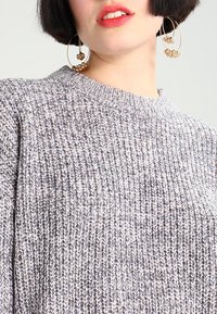 Szary sweter dzianinowy z teksturowanej tkaniny o prążkowanym wzorze, okrągłym dekolcie i opadających ramionach. Uzupełniony dużymi kolczykami w kształcie kół.