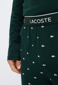 Pantalones de pijama de algodón verdes con logos blancos de Lacoste, con una cinturilla negra que lleva "LACOSTE" en blanco. Textura suave, ajuste casual.