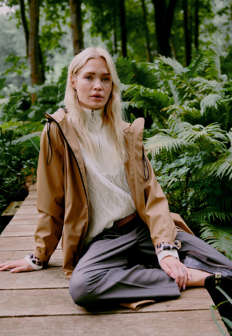 Veste imperméable marron avec capuche à cordon, pull en tricot blanc, pantalon large gris et chaussures noires, assis sur un passage en bois dans la verdure.