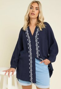 Camicia di cotone blu navy con maniche lunghe, caratterizzata da motivi ricamati bianchi sul davanti e una vestibilità ampia. Abbinata a pantaloni corti in denim.