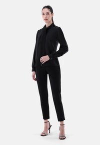 Black blouse with a draped collar, long sleeves, and button details, paired with slim-fit black pants and black strappy high heels.