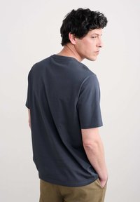 Camiseta de algodón gris oscuro con mangas cortas y cuello redondo, vista desde la parte de atrás, con las manos en los bolsillos.