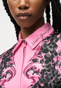 Κοντινό πλάνο ενός ατόμου που φοράει ένα ροζ και μαύρο πουκάμισο με σχέδιο, με γιακά που φέρει το κείμενο "Versace Jeans Couture" στον γιακά.