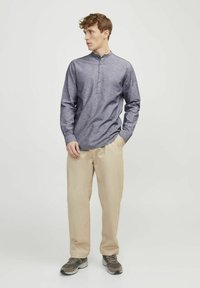 Camicia a maniche lunghe con bottoni in tessuto blu scuro, dotata di colletto alla mandarina e quattro bottoni frontali, abbinata a pantaloni beige e sneakers grigie.