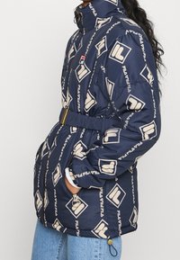 Granatowa kurtka puchowa z wzorem logo, wysokim kołnierzem, elastycznym paskiem w talii, kieszeniami na zamek i złotymi akcentami do regulacji.