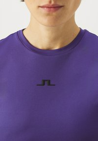 Violet sportinis marškinėliai su apvaliu iškirpimu ir juodu logotipo akcentu centre. Lygus paviršius, prigludusi konstrukcija.