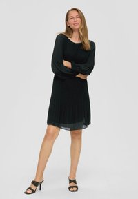 Schwarzes plissiertes Kleid mit langen Ärmeln, Rundhalsausschnitt und fließendem Saum. Getragen mit schwarzen High-Heels. Glatte Textur.