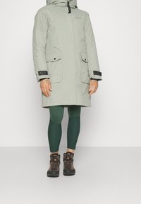 Parka gris clair avec capuche, comprenant des accents noirs, deux grandes poches avant et des poignets réglables, portée avec des leggings verts et des bottes.