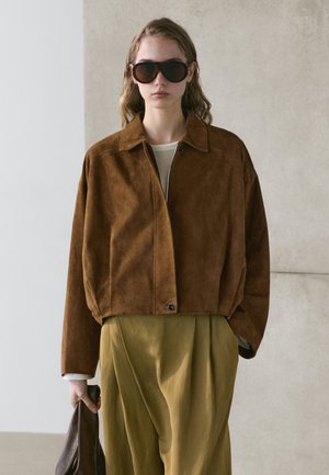 Kobieta ubrana w dużą brązową zamszową kurtkę i oliwkowe spodnie, trzymająca w jednej ręce brązową skórzaną torbę oraz nosząca duże ciemne okulary przeciwsłoneczne.