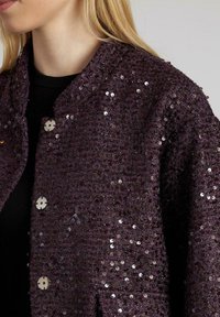 Donna che indossa una giacca texturizzata in paillettes viola scuro con grandi bottoni argento sopra una maglietta nera, mostrata dalla zona del collo fino a metà torace.