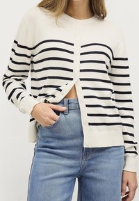 Cardigan rayé en crème avec des rayures horizontales bleu marine, doté d'un col rond, de cinq boutons et de poignets côtelés. Assorti à un jean bleu clair.