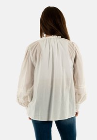 Blusa bianca con maniche a sbuffo, delicata bordatura in pizzo ai polsini e dettagli arricciati sul retro, realizzata in tessuto leggero.